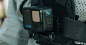 A GoPro Hero 13 Black action camera is mounted on a chest strap, attached to the mesh fabric of a gray jacket. The camera has a rectangular lens and a small screen on the front.