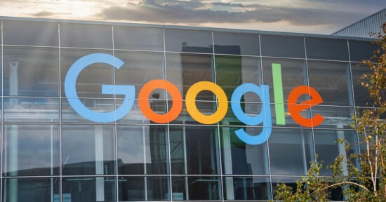 A large sign displaying the word "Google" in colorful letters is mounted on the glass facade of a modern building. The background includes a sky with clouds and some sunlight. A tree with green leaves is visible in the bottom right corner.