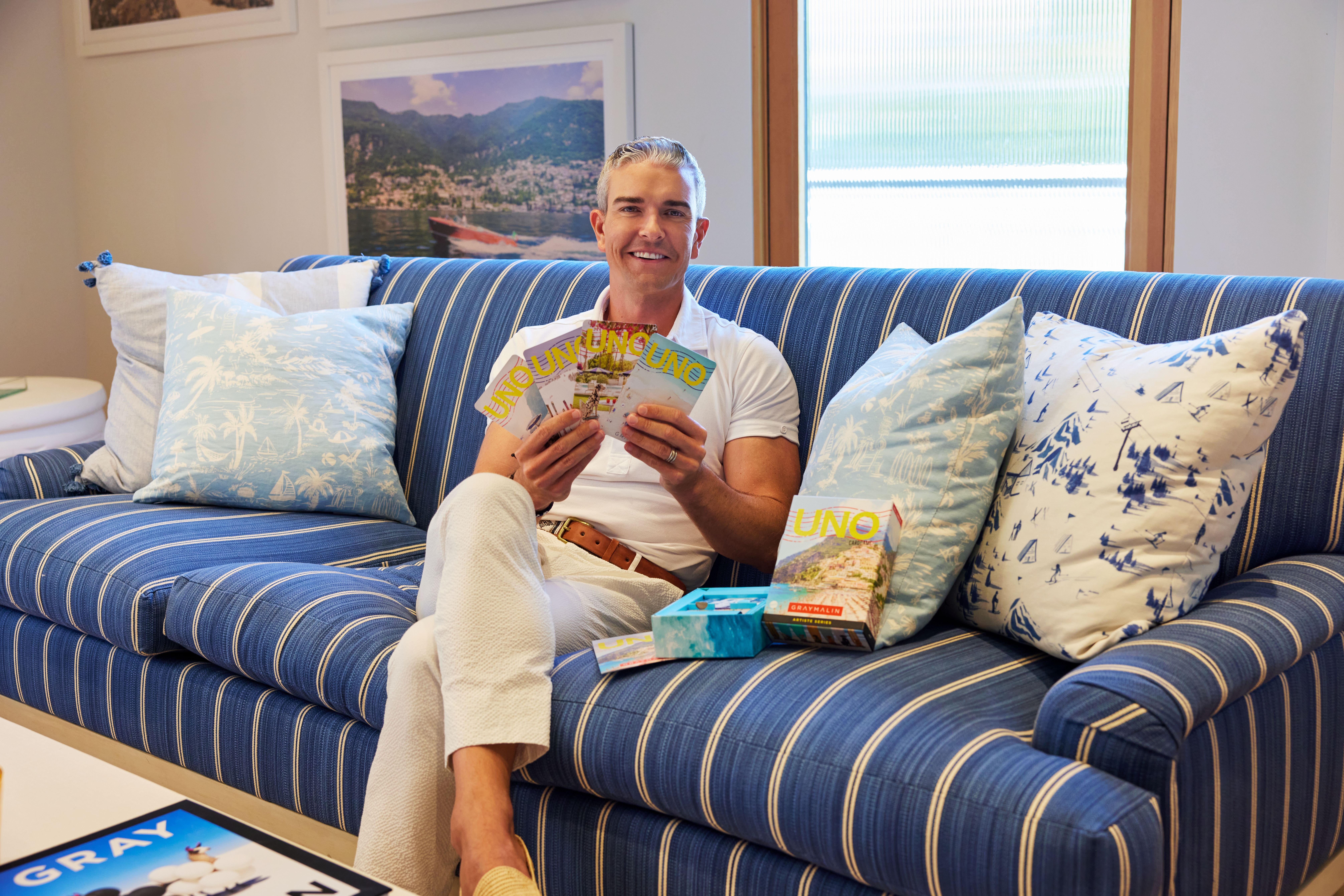 A smiling man sits on a blue striped sofa holding several UNO cards. He is dressed in a white shirt and pants. Visible around him are cushions with blue and white patterns, and a box of UNO cards placed on the sofa. In the background are framed pictures on the wall.