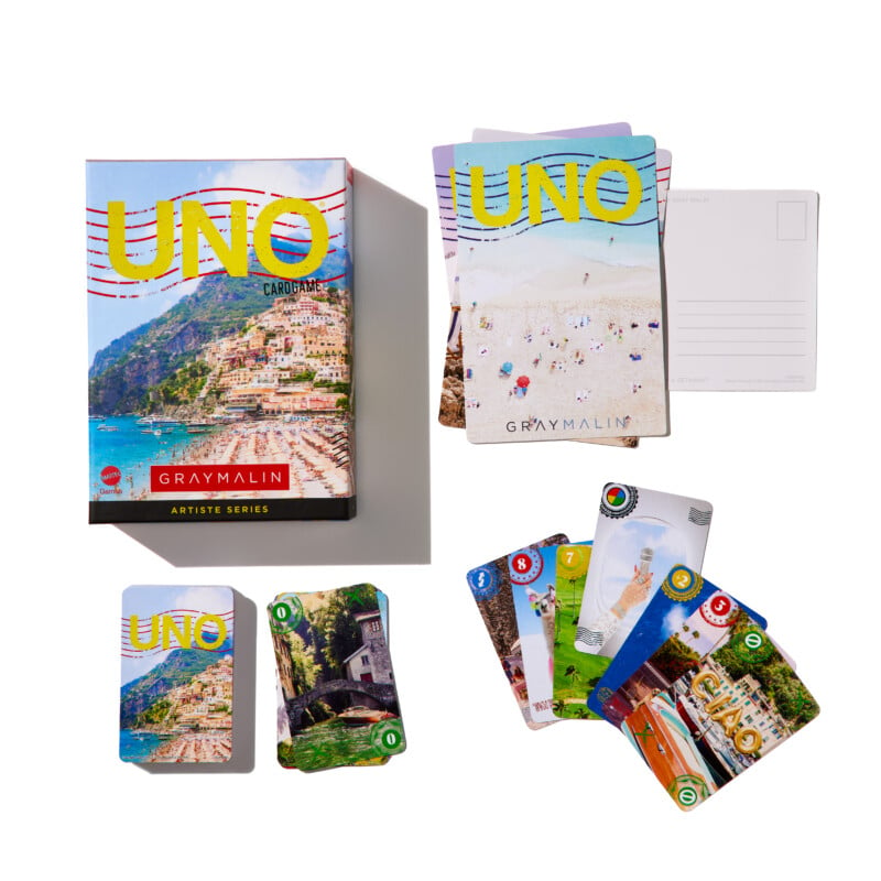 A Gray Malin-themed UNO card game is shown. It includes a box and several cards decorated with colorful, scenic images. The cards are fanned out, displaying different landscapes and scenes. The box features a vibrant photo and the title "UNO" and "Gray Malin.