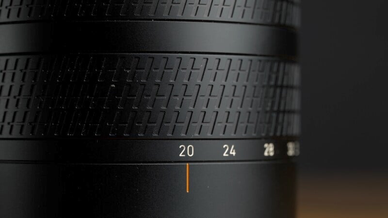 Close-up of a black camera lens showing detailed texture and markings, with numbers "20" and "24" clearly visible on the barrel. The lens is set against a dark background. An orange line aligns with the marking "20.