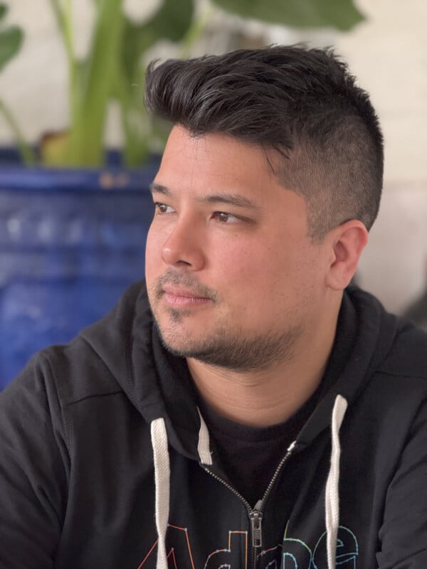 A man with short dark hair and a trimmed beard wearing a black hoodie is looking to the side. There is a large blue ceramic pot with greenery in the background. The image is well-lit with natural light.