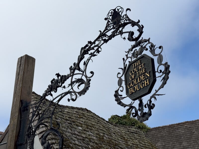 An ornate wrought-iron sign with intricate designs hangs above a shingled roof. The sign reads, "The Court of the Golden Bough." The sky in the background is partially cloudy.