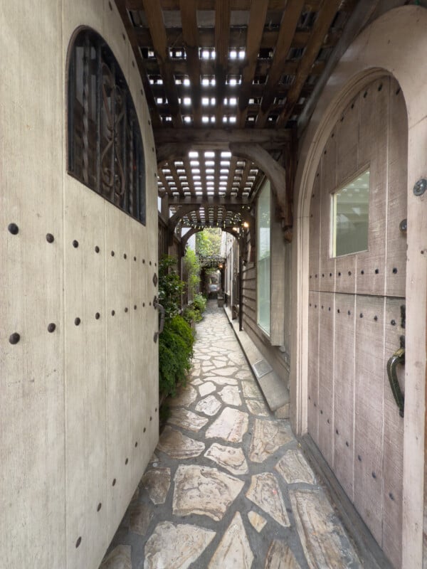 A narrow, covered pathway with a stone floor leading through a shaded area framed by wooden doors and trellises. Green foliage lines parts of the path, and the wooden structures have a rustic, weathered appearance. Light filters through the overhead lattice.