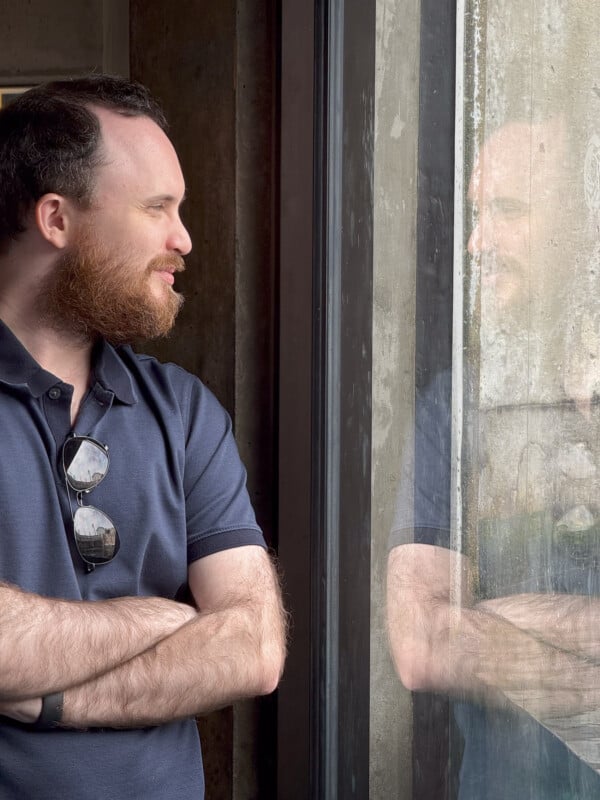 A man with a beard and glasses hanging from the collar of his dark polo shirt is standing with his arms crossed, looking out of a window. His reflection is visible in the glass, giving the impression that he is facing himself.