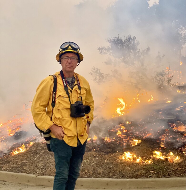 A person stands in front of a wildfire, wearing a yellow fire-resistant jacket, helmet, and goggles. They have a camera around their neck and another camera slung over their shoulder. Smoke and flames are visible in the background.