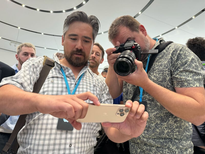 Two men are focused on a smartphone, which one man is holding, while the other is taking a close-up photo with a professional camera. Several people are in the background, and they are all in a well-lit room with modern design elements.