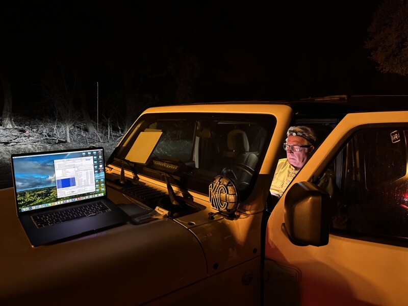 A man sits in the driver's seat of an off-road vehicle at night, illuminated by a light. A laptop with a landscape wallpaper and multiple open applications is placed on the vehicle's hood. The background is dark, with trees and bushes barely visible.