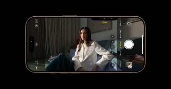 A smartphone display shows a camera app interface capturing a woman who is sitting on a couch with her arms crossed. The woman has long hair and is dressed in a white top and green pants. Various camera settings are visible on the screen.