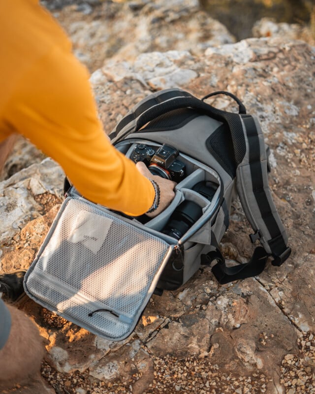 A person in a long-sleeve yellow shirt is accessing a grey camera backpack placed on rocky terrain. The backpack is open, revealing compartments containing a camera and lens. The person has their hand inside the backpack, reaching for the equipment.