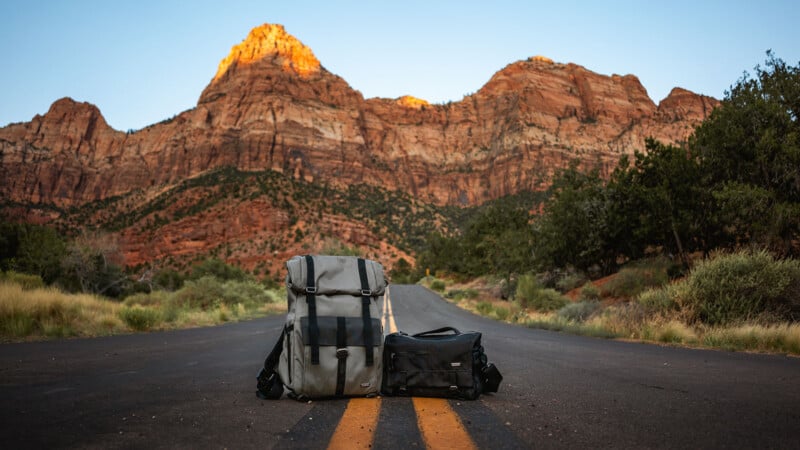 Two backpacks rest on a road's yellow median line, framed by towering red rock formations and a bright blue sky in the background. Green shrubs and trees line the sides of the road, creating a picturesque and serene outdoor scene.