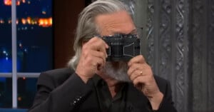 A person with gray hair and a beard is holding an old-fashioned camera up to their face as if taking a photo. The background includes a window with city lights and an ornate patterned wall. The person is wearing glasses and a dark-colored shirt or jacket.