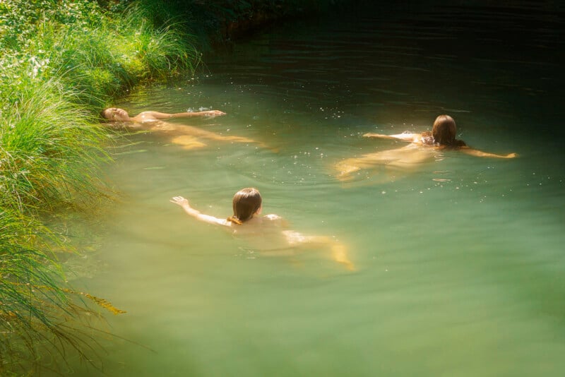 Three people float leisurely in a serene, natural pond. Their bodies are partially submerged in the calm water, surrounded by lush green grass and vegetation. Sunlight filters through the trees, casting gentle shadows and illuminating the scene.