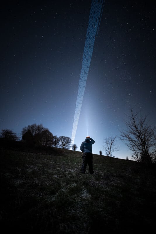 A person stands in a grassy field at night, looking up at the sky. The clear night sky is adorned with stars and a long, linear streak of light that stretches diagonally from the horizon to the top of the image. Trees silhouetted against the sky line the horizon.