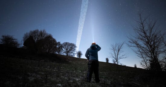 A person in a blue jacket stands on a grassy field at night, looking up at the starry sky with a bright light beam extending from their headlamp into the sky. Silhouetted trees are visible in the background.