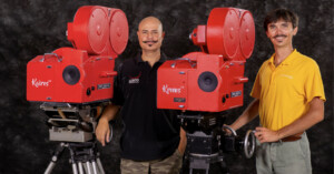 Two men stand beside two vintage red movie cameras on tripods. The man on the left wears a black shirt with logos, while the man on the right wears a yellow polo shirt. Both smile and look into the camera, with a dark backdrop behind them.