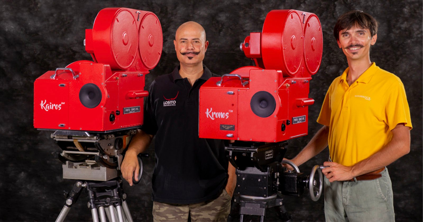 Two men stand beside two vintage red movie cameras on tripods. The man on the left wears a black shirt with logos, while the man on the right wears a yellow polo shirt. Both smile and look into the camera, with a dark backdrop behind them.