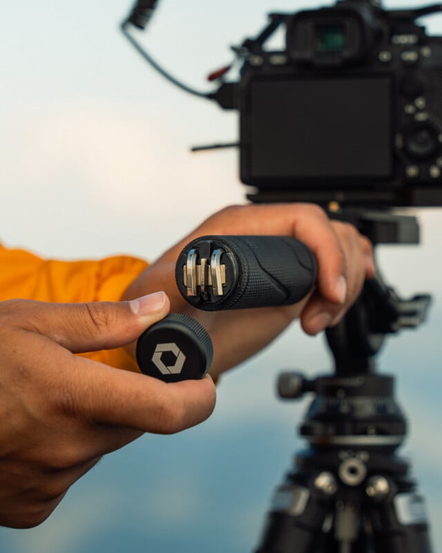 A person holds a cylindrical device with multiple metallic connectors inside, with the cap removed. The device is being demonstrated in front of a camera mounted on a tripod. The background is out of focus.