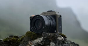 A Hasselblad camera is positioned on a moss-covered rock outdoors against a foggy, mountainous backdrop. The camera's design is sleek and modern, featuring a large lens and branding that reads "Hasselblad." The setting gives a rugged and natural feel.