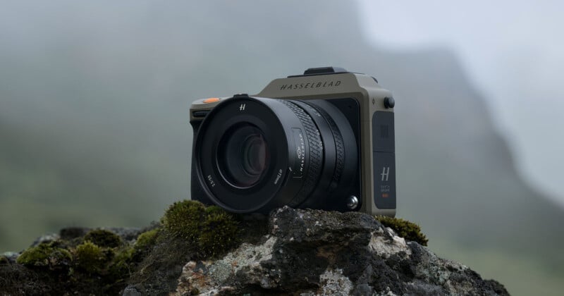 A Hasselblad camera is positioned on a moss-covered rock outdoors against a foggy, mountainous backdrop. The camera's design is sleek and modern, featuring a large lens and branding that reads "Hasselblad." The setting gives a rugged and natural feel.