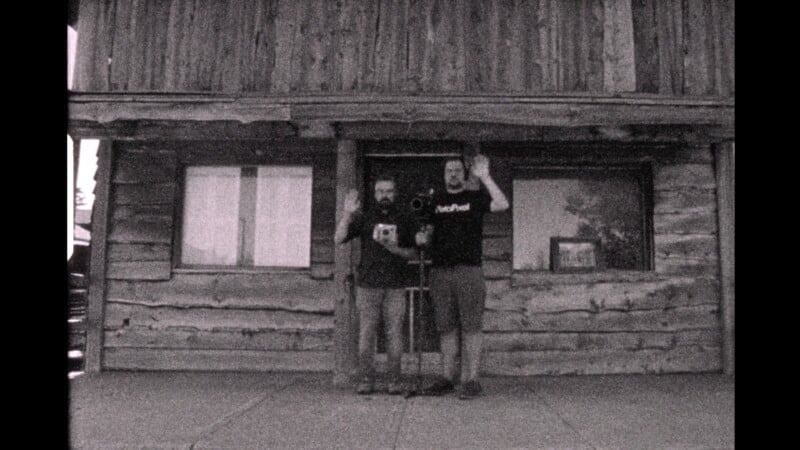 A grainy, black and white photo of two people standing on a sidewalk in front of a rustic wooden building. One person is holding a vintage camera, and the other is standing beside them with an arm raised in greeting. They are both looking towards the camera.