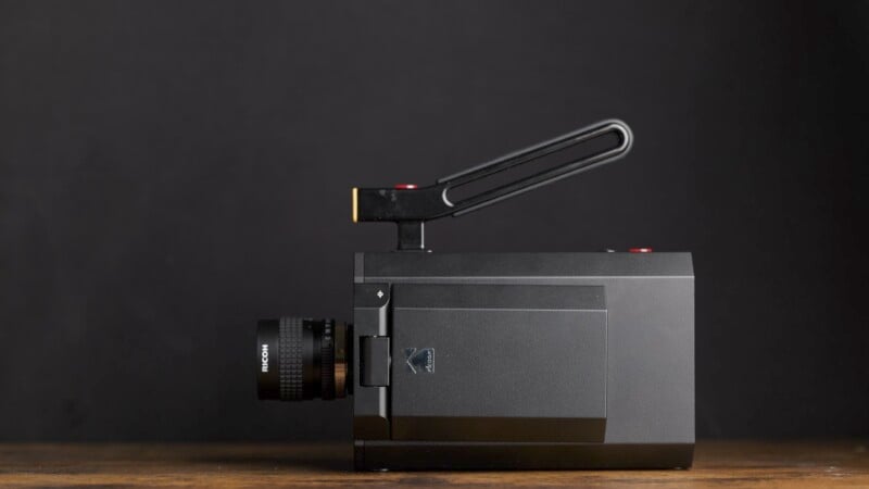 A vintage black video camera with a side handle is placed on a wooden surface against a dark background. The camera features a prominent lens and several buttons, giving it a classic and professional appearance.