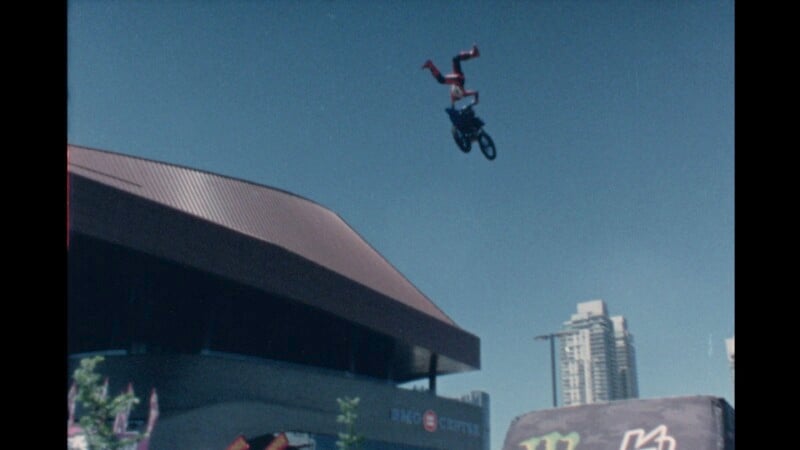 A person performs an aerial stunt on a BMX bike, releasing the bike while upside down in mid-air. They are high above the ground near a large building with a brown, curved roof. Skyscrapers and a clear blue sky form the background.