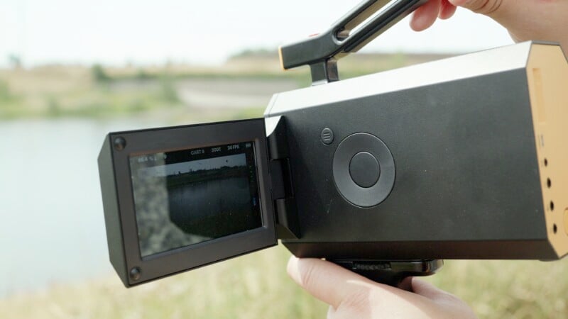 A person is holding a black camcorder with an open flip-out screen, recording a scene outdoors near a body of water. The background appears slightly blurred, focusing on the camcorder and screen, which shows a preview of the recording.