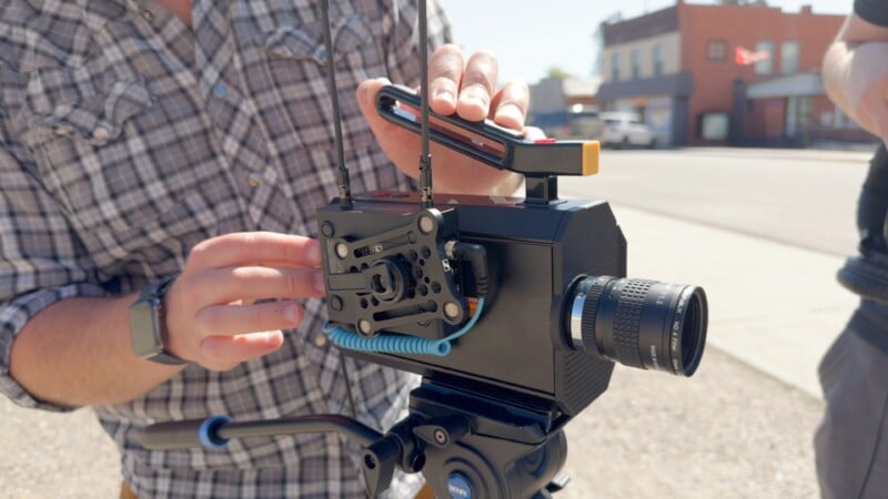 A person dressed in a plaid shirt is adjusting a professional video camera mounted on a tripod outdoors. The camera has various attachments, including a microphone and a manual focus lens. The background shows a street with buildings and a clear sky.