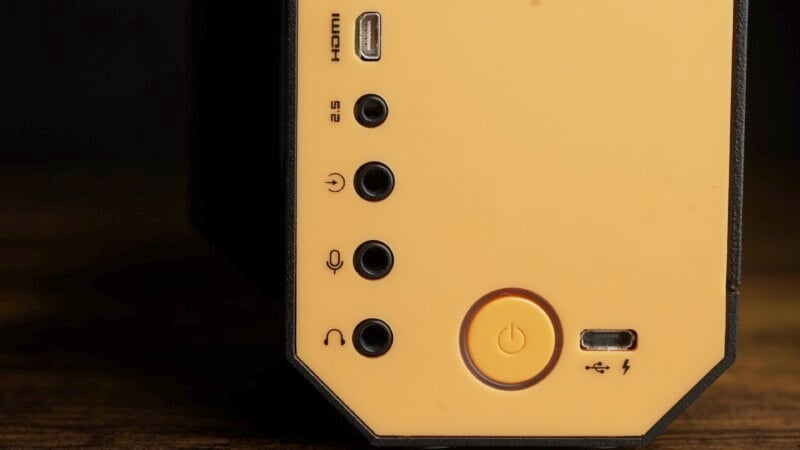 Close-up view of a panel featuring a power button, headphone jack, microphone input, 2.5mm port, HDMI port, and a USB port, with a warm yellow background and subtle lighting. The arrangement indicates typical connectivity options for an electronic device.