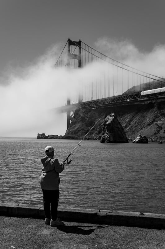 A black-and-white image shows a person fishing at the water's edge, with the Golden Gate Bridge partially shrouded in fog in the background. The fisher wears a cap and hoodie, standing near a rocky shoreline as the iconic bridge towers behind.