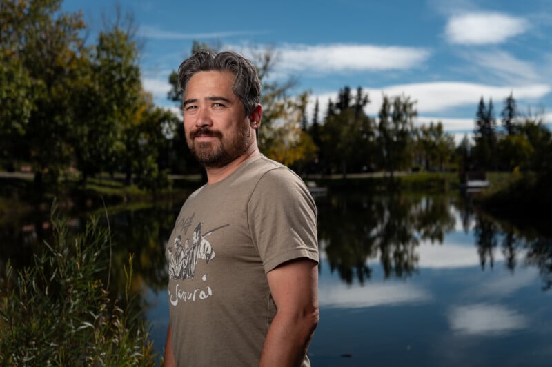 A man with gray-streaked hair and a beard stands outdoors near a reflective pond. He is wearing a beige t-shirt with a graphic design. The background shows trees, greenery, and a partly cloudy blue sky.