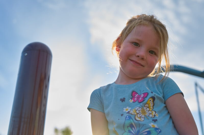 A young girl with light hair smiles at the camera. She is wearing a blue t-shirt with colorful butterfly prints. The background shows a blurred outdoor scene with a blue sky and some playground equipment. The lighting creates a soft, warm glow around her.