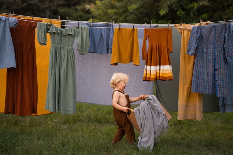 A young child with blonde hair, shirtless and wearing brown overalls, stands on the grass, holding a grey blanket. Behind the child, a clothesline with various colorful garments is set up outdoors against a backdrop of trees.