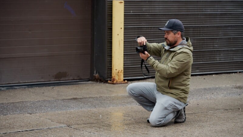 A person wearing a green jacket and a cap kneels on a wet pavement while adjusting a camera. They are positioned in front of a closed metal garage door. The scene appears to be overcast, with muted colors and a calm atmosphere.