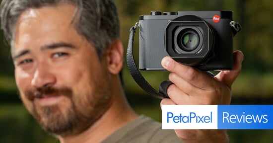 A person holding a camera close to the lens, slightly out of focus. The background is blurred greenery. There's a logo for PetaPixel Reviews in the lower right corner.