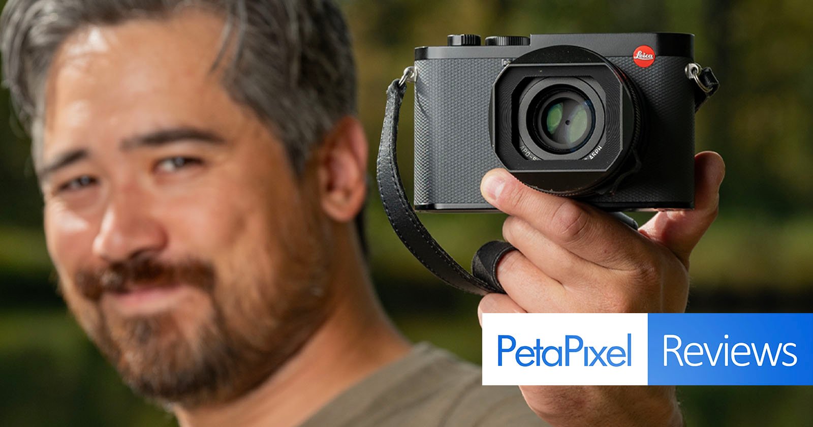 A person holding a camera close to the lens, slightly out of focus. The background is blurred greenery. There's a logo for PetaPixel Reviews in the lower right corner.