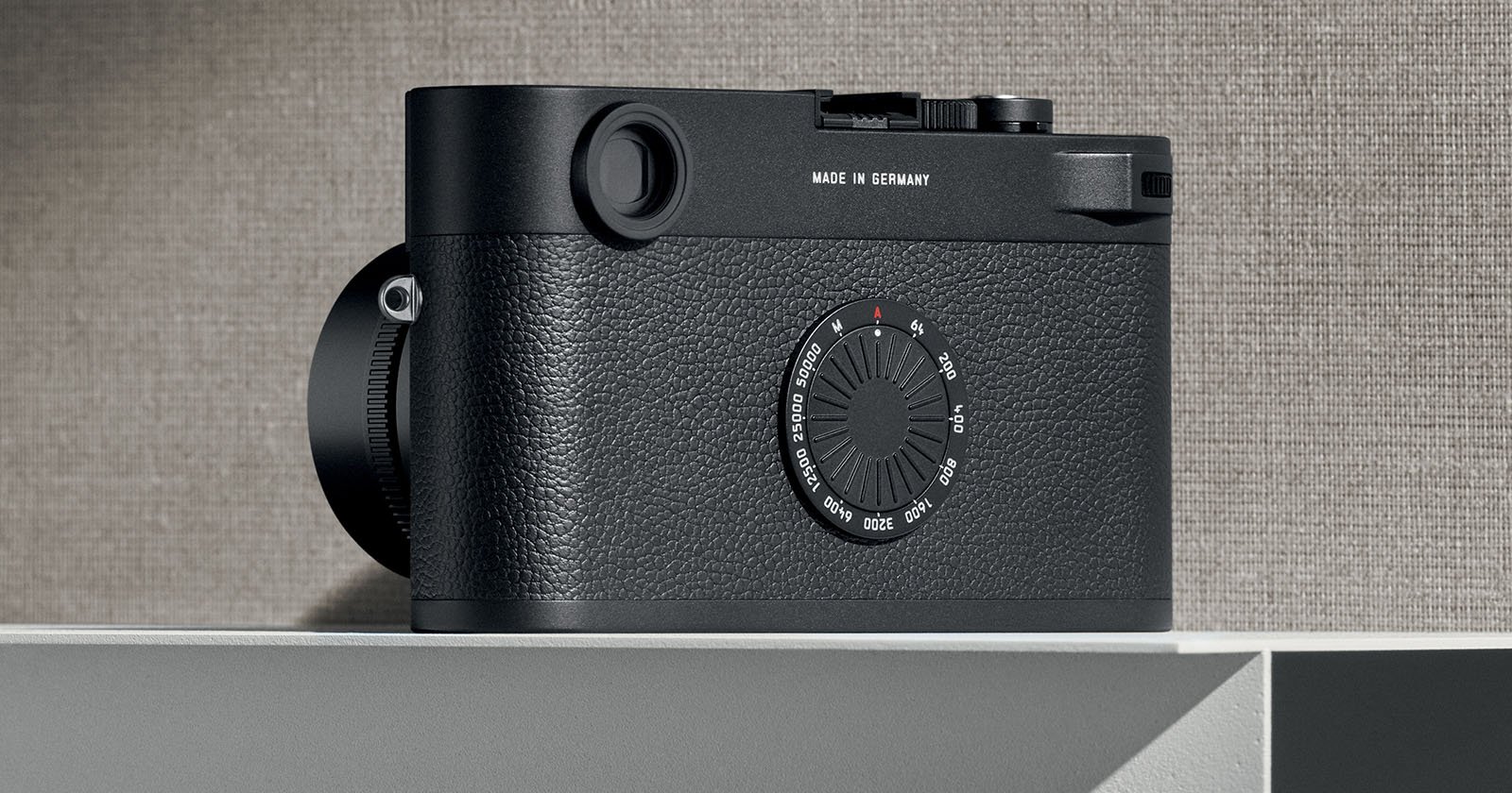 A black, compact digital camera with a textured grip on a white surface. The camera's design features a lens on the left side and various dials on the top. The text "MADE IN GERMANY" is visible on the top right section. The background is a textured gray wall.