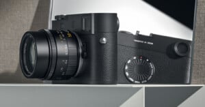 A black digital Leica camera with a lens, positioned on a white shelf next to a grey mirror. The camera has a sleek, modern design with a textured grip. Behind the camera, its reflection is visible in the mirror. The background is minimalistic.