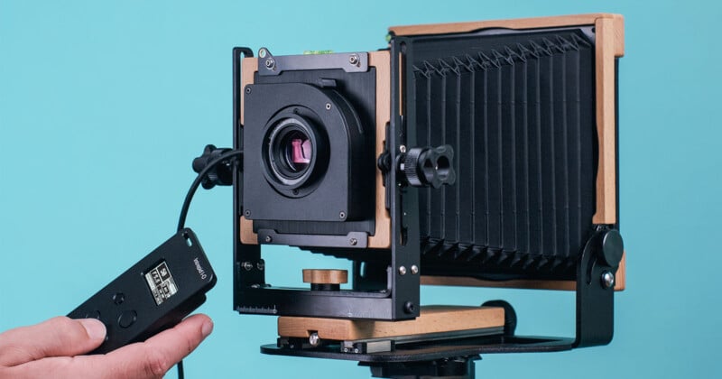 A close-up of a wooden large format camera with bellows, mounted on a tripod against a blue background. A person is holding a remote control attached to the camera, preparing to take a photograph.