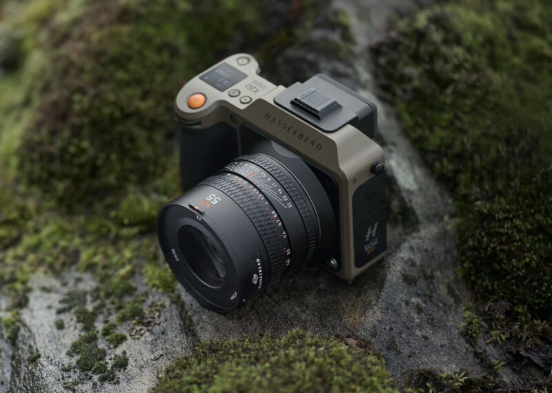 A Hasselblad digital camera with a 55mm lens is placed on a moss-covered rocky surface. The camera has a rugged, matte green and black exterior, designed for outdoor use.