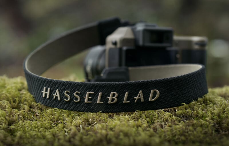 A close-up of a black camera strap with the word "Hasselblad" embroidered in white, lying on a patch of green moss. Blurred background shows a camera attached to the strap.