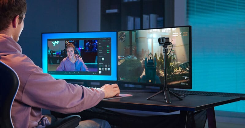 A person is sitting at a desk playing a video game on one monitor and streaming or video chatting on another. They wear a hoodie and use a keyboard and mouse. A webcam on a tripod is positioned between the monitors. The room has a modern, tech-focused setup.