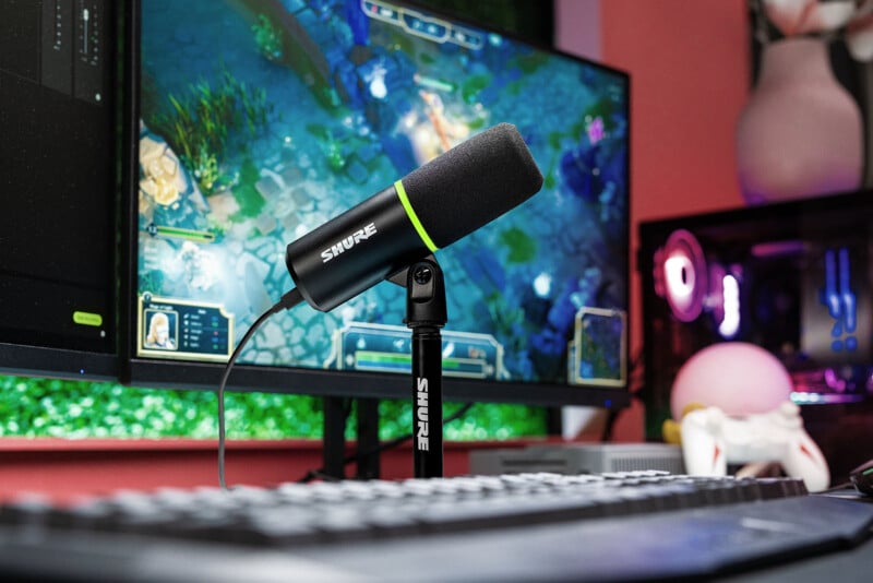 A desktop setup featuring a Shure microphone in the foreground, positioned in front of a keyboard. Behind the microphone, there are two monitors displaying a video game, and to the right, a gaming PC with colorful LED lighting is partially visible.