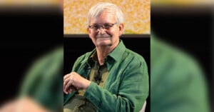 An elderly person with white hair, glasses, and a green jacket smiles while sitting. The background features a blurry yellow and white pattern.