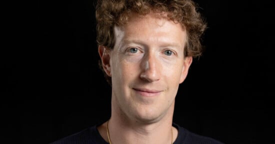 A person with curly brown hair, light skin, and blue eyes is seen in a close-up portrait against a black background. They are wearing a dark-colored shirt and a subtle smile is visible on their face.