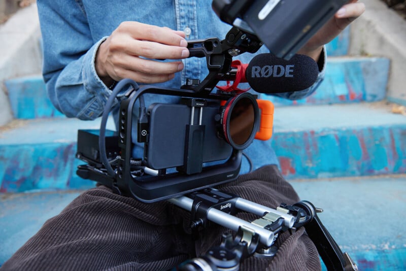 A person wearing a blue denim shirt sits on stairs, assembling a camera rig with a mounted microphone labeled "RØDE." The camera is attached to a stabilizing frame, and the person is adjusting components while resting the setup on their lap.