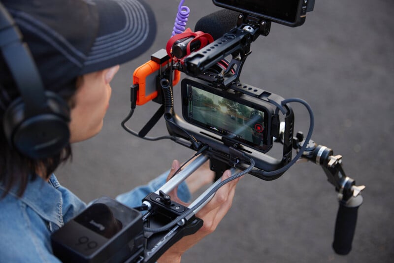 A person wearing headphones and a cap operates a complex camera rig outdoors. The rig includes a smartphone, various attachments, and stabilization equipment. The smartphone displays a video recording in progress.