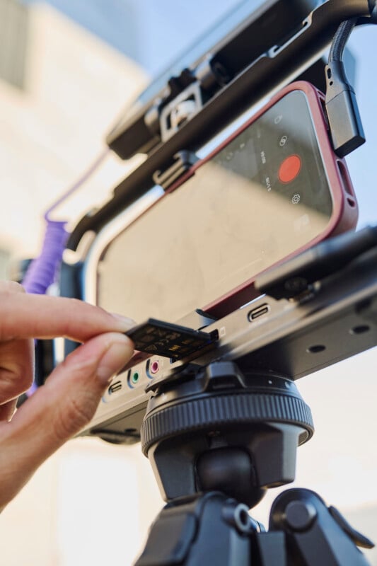 A close-up of a hand inserting a memory card into a small video camera setup mounted on a tripod. The camera setup includes a smartphone, attached horizontally within a rig, with recording software visible on the screen.