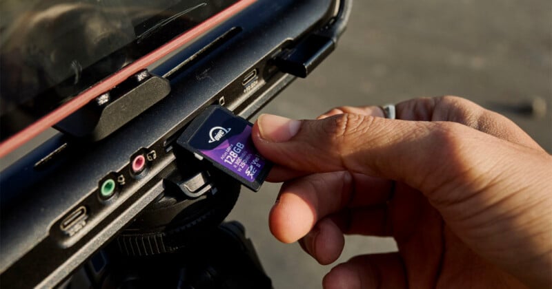 A close-up image of a person's hand inserting a purple 128GB SD card into a slot on the side of a camera or similar device. The person's fingers are holding the edge of the card, and different input ports are visible around the slot area.